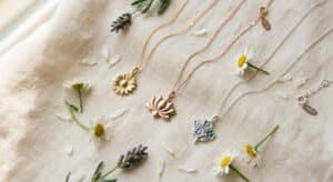 A flat lay of three flower necklaces on cream linen — a gold daisy flower necklace, a rose gold lotus flower necklace, and a sterling silver forget-me-not flower necklace, styled with fresh daisies and lavender